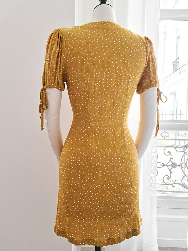 A short summer dress in ochre yellow with white polka dots is presented on a couture mannequin in a bright white room. The dress is seen from the back.