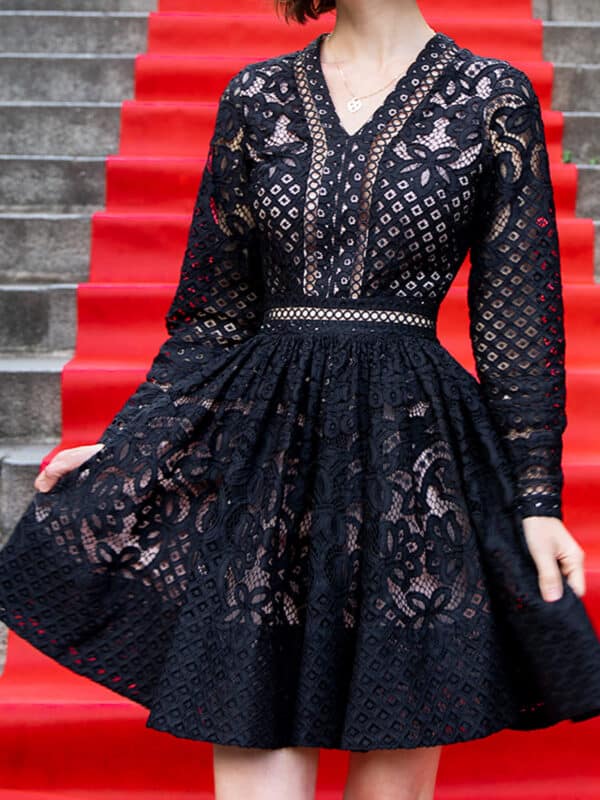 A young woman wears a black lace dress with V neck, fitted bodice and lace details on the bodice. The skirt is very full and she holds it in her hand while walking down a staircase covered with a red carpet.