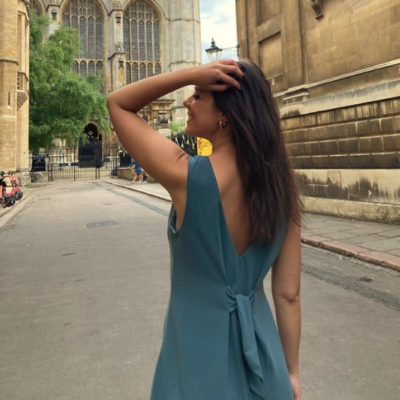 A young brunette woman wears the Vega summer dress pattern in a blue green crepe. She is tossing her hair to the side, walking in the street.