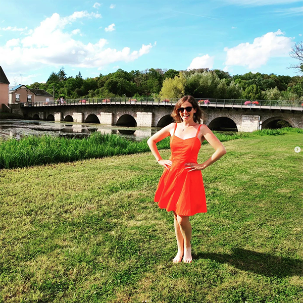 Sirius summer dress sewing pattern in a bright orange fabric, modeled by a young woman in front of a bridge in the countryside. She put her hands on her hips and is smiling to the camera while a gentle wind shows the fullness of the skirt.