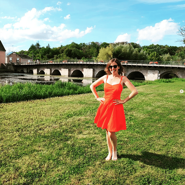 Sirius summer dress sewing pattern in a bright orange fabric, modeled by a young woman in front of a bridge in the countryside. She put her hands on her hips and is smiling to the camera while a gentle wind shows the fullness of the skirt.