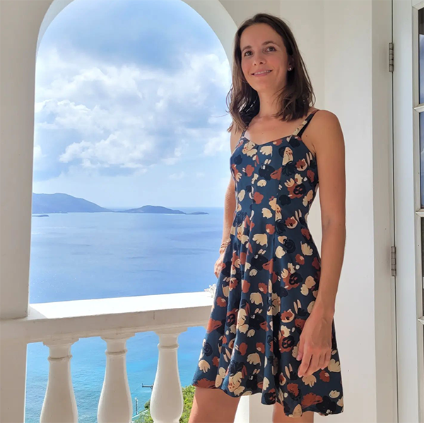 Sirius slip dress sewing pattern in a printed viscose, modeled by a young woman with shoulder-length hair, standing in front of a window by the sea
