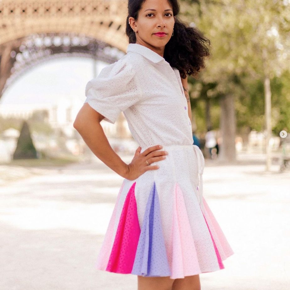A young black woman wears a white shirt dress with a godet skirt. The godets are made in contrasting colors, in a cameo of pink and purple.