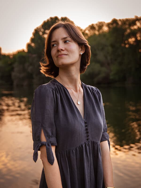 A young brunette woman with short hair is wearing the Pleiades 1 dress in a black tercel crepe, in the tied sleeve version. She is standing by a river, and the sun is setting in the background, creating a gold halo around her.