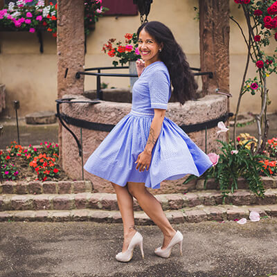 A woman with long black curly hair is twirling in a lavender blue dress with very full skirt. She is smiling and dancing in a garden of roses. The dress is made of an eyelet lace and the bodice is adorned with a white eyelet detail.