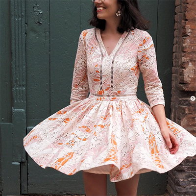 A young brunette woman wears the Aquarius dress sewing pattern as her wedding guest outfit. She made the dress in a pastel pink and orange eyelet cotton. She is twirling around, showing the very full skirt.