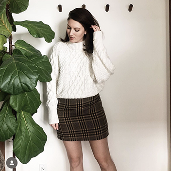 A young brunette woman with long hair is wearing a brown tartan mini skirt with a white aran sweater.