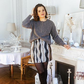 A young brunette woman wears a glittery silver and gold mini skirt with a grey loose sweater. She stands in a brightly lit living room, decorated for the holidays.