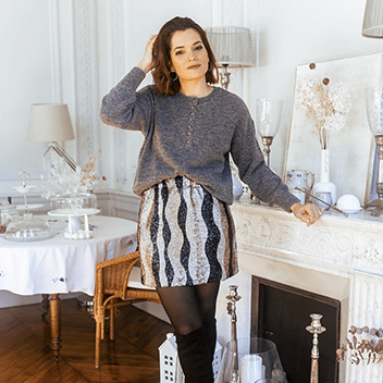 A young brunette woman wears a glittery silver and gold mini skirt with a grey loose sweater. She stands in a brightly lit living room, decorated for the holidays.