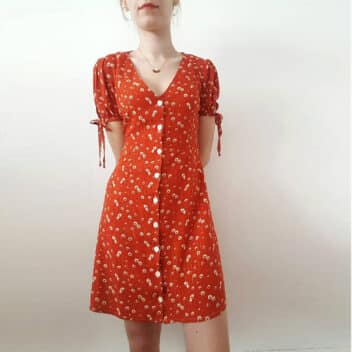 A young woman wears a red dress with tiny floral pattern that hugs her curves. The dress reaches mid-thigh, is buttoned at the front and has short, gathered and knotted sleeves.